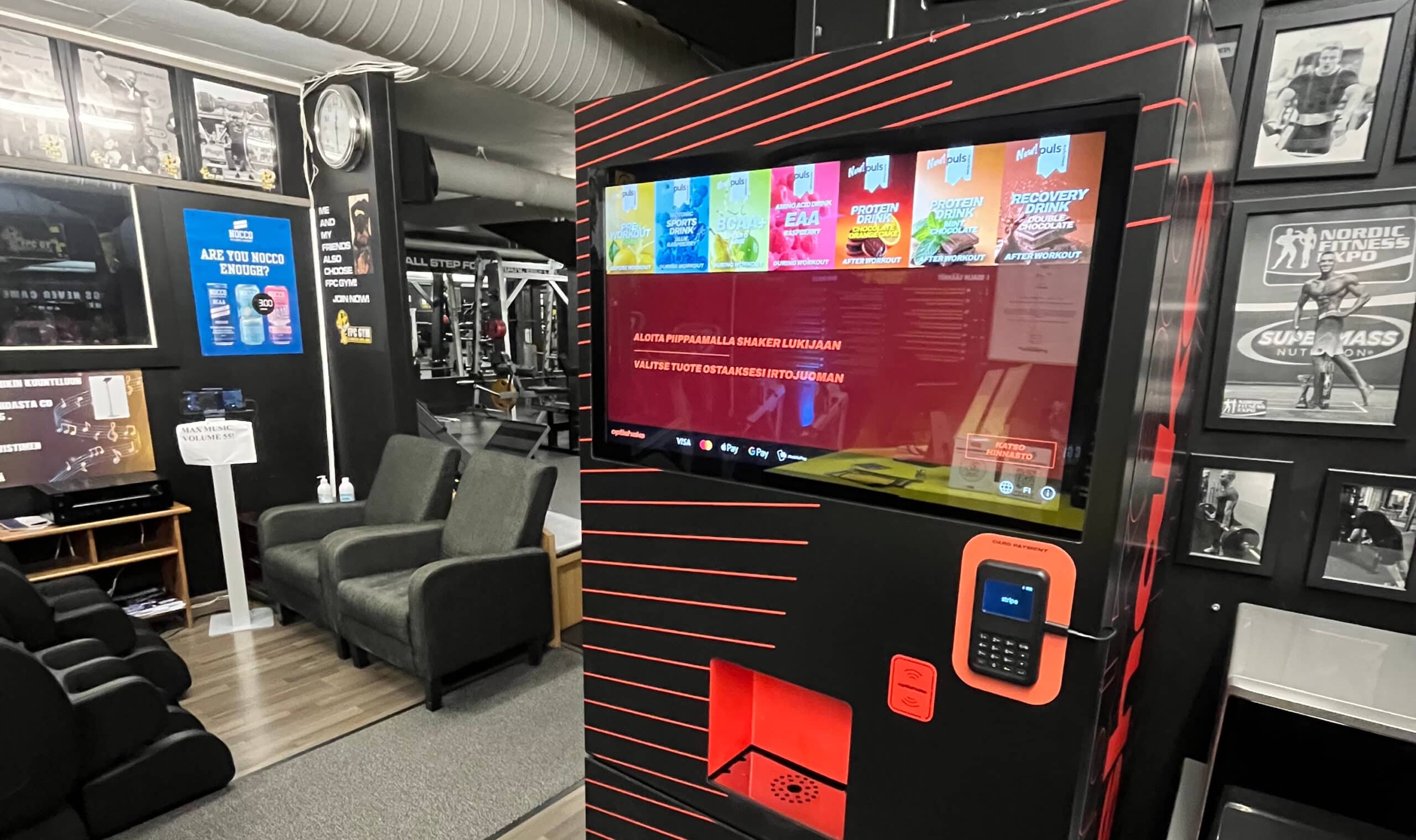 Automaatin kuva FPC GYM:ssä Hamina, jossa uusi juomakone tarjoaa proteiinijuomia ja palautusjuomia treenaajille. Moderni ja helppokäyttöinen laite sijaitsee kuntosalin sisällä, edistäen hyvinvointia j.